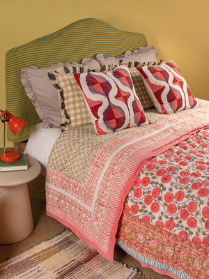A cozy bed features a colorful striped headboard, patterned pillows, and the Aiko Cotton Quilt by Ian Snow Ltd with geometric floral design. An orange lamp and books sit on the side table against a yellow wall with a woven rug below.