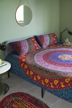 A neatly made bed showcases the vibrant Sanganeri Cotton Duvet Set by Ian Snow Ltd and matching pillows against pale green walls. A round mirror, bedside table with books, and two black lamps complete the look.