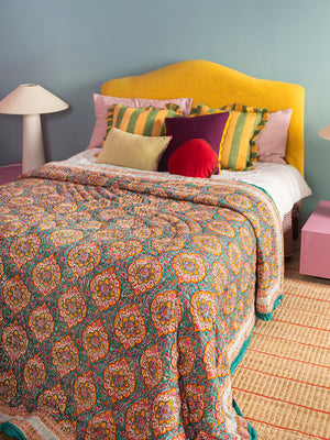 A cozy bedroom features a bed with a yellow headboard, an Ian Snow Ltd Jasmine Block Print Cotton Quilt, striped and solid pillows, pink side tables, white lamps, and a woven rug on the floor against a light blue wall.