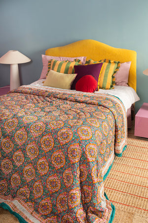 A cozy bedroom features a bed with a yellow headboard, an Ian Snow Ltd Jasmine Block Print Cotton Quilt, striped and solid pillows, pink side tables, white lamps, and a woven rug on the floor against a light blue wall.
