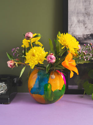 A Marble Finish Glass Hurricane Vase by Ian Snow Ltd sits on a purple surface, with a black rotary phone to its left and part of a framed picture in the background against a green wall.