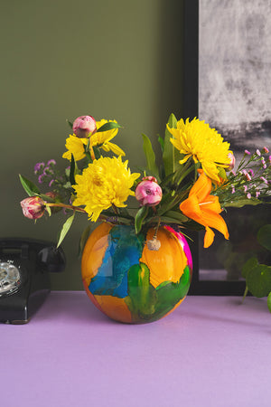 A Marble Finish Glass Hurricane Vase by Ian Snow Ltd sits on a purple surface, with a black rotary phone to its left and part of a framed picture in the background against a green wall.