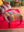 The Ian Snow Ltd Striped Velvet Footstool, featuring vibrant stripes and wooden legs, sits on a red rug by a matching armchair. A wooden side table and a green wall are visible in the background.