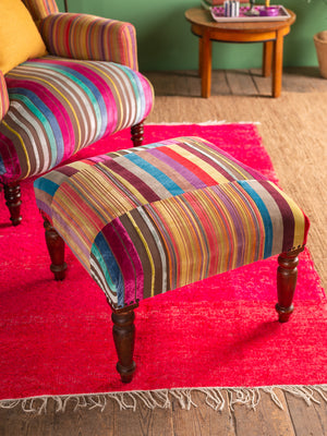 The Ian Snow Ltd Striped Velvet Footstool, featuring vibrant stripes and wooden legs, sits on a red rug by a matching armchair. A wooden side table and a green wall are visible in the background.