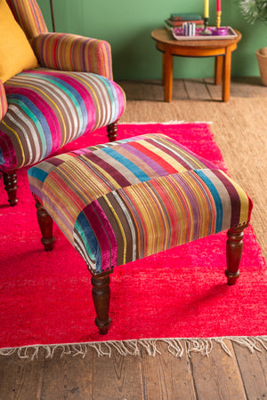 The Ian Snow Ltd Striped Velvet Footstool, featuring vibrant stripes and wooden legs, sits on a red rug by a matching armchair. A wooden side table and a green wall are visible in the background.
