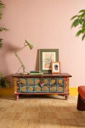 A Blue Painted Trunk made from New and Reclaimed Wood with a Metal Trim by Ian Snow Ltd sits against a pink wall, topped with two framed artworks, a pale green lamp, and stacked books. Plants and a rug add warmth to the cozy scene.