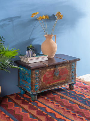 The Ian Snow Ltd Red and Blue Reclaimed Wooden Chest with Brass Trim sits against a blue wall, topped with stacked books, a terracotta vase of dried yellow flowers, and a small plant. A red patterned rug adds to the cozy atmosphere.