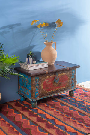 The Ian Snow Ltd Red and Blue Reclaimed Wooden Chest with Brass Trim sits against a blue wall, topped with stacked books, a terracotta vase of dried yellow flowers, and a small plant. A red patterned rug adds to the cozy atmosphere.