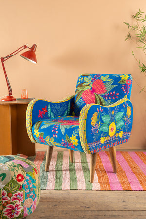 The Cholena Embroidered Armchair by Ian Snow Ltd, featuring vivid floral patterns, sits on a striped rug beside a wooden side table with a red lamp, set against a peach wall and leafy plant in the corner.