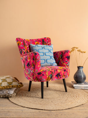 The Cerise Bird of Paradise Velvet Armchair by Ian Snow Ltd features a vibrant pink hue and floral design. It sits on a round woven rug with a blue patterned cushion, decorative pillows, stacked books, and dried flowers by a peach wall.