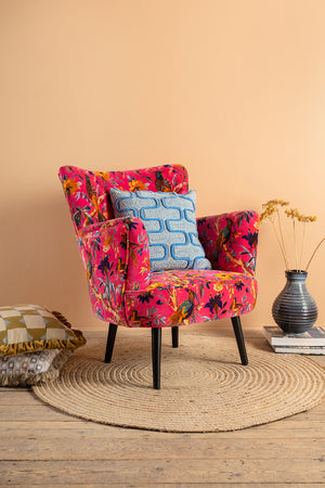 The Cerise Bird of Paradise Velvet Armchair by Ian Snow Ltd features a vibrant pink hue and floral design. It sits on a round woven rug with a blue patterned cushion, decorative pillows, stacked books, and dried flowers by a peach wall.