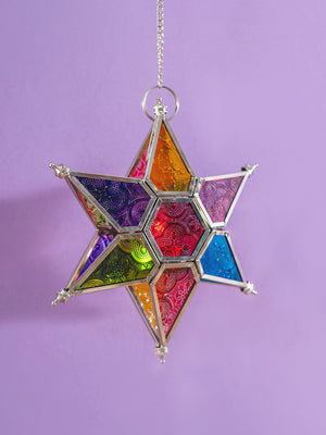 The Multi Coloured Iron & Glass Hanging Star Lantern by Ian Snow Ltd features intricate patterns and vibrant panels, suspended from a chain against a light purple background.