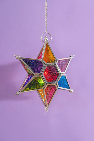 The Multi Coloured Iron & Glass Hanging Star Lantern by Ian Snow Ltd features intricate patterns and vibrant panels, suspended from a chain against a light purple background.