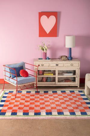 Living room with a colorful checkered rug, red chair, and pink wall with a heart-shaped painting.