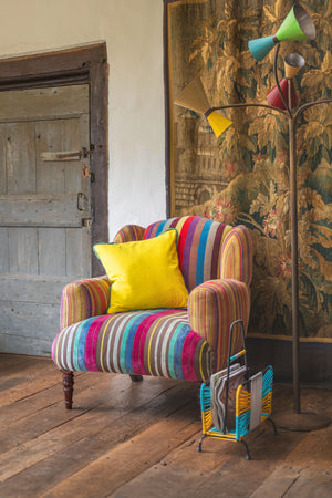 The Ian Snow Ltd Striped Velvet Armchair with a yellow pillow sits on a wooden floor beside a retro floor lamp and magazine rack, set against a rustic door and tapestry.