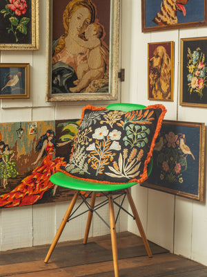 A cushion cover with a jet black background featuring intricate embroidered plant life, displayed on a chair against a backdrop of various framed artworks.