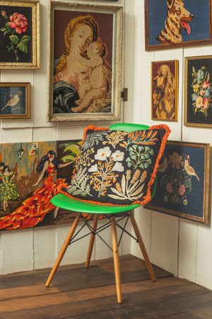 A cushion cover with a jet black background featuring intricate embroidered plant life, displayed on a chair against a backdrop of various framed artworks.