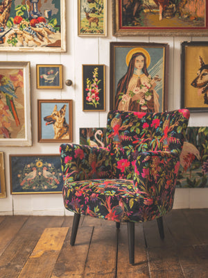 The Black Bird of Paradise Velvet Chair by Ian Snow Ltd, featuring a vibrant floral and bird pattern, sits on a wooden floor amid a gallery wall of framed art with animals, flowers, and a haloed woman holding a lamb.