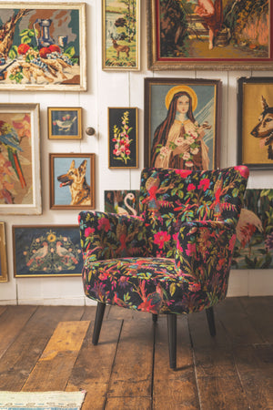 The Black Bird of Paradise Velvet Chair by Ian Snow Ltd, featuring a vibrant floral and bird pattern, sits on a wooden floor amid a gallery wall of framed art with animals, flowers, and a haloed woman holding a lamb.
