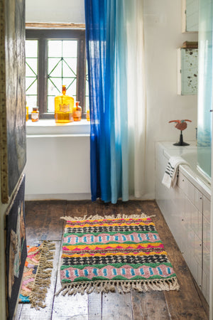 A cozy bathroom with wooden floors, white walls, blue-curtained window, amber bottles on the sill, a towel by the tub, and the Ian Snow Ltd Palazzo Geometric 100% Recycled Small Rug adds stylish comfort.