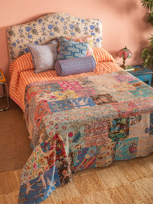 A bed with a floral headboard, orange striped sheets, and the Lola Patchwork Kantha Bed Throw by Ian Snow Ltd. Four patterned pillows are at the head, with a vintage pink lamp and leafy plant on a blue nightstand nearby.