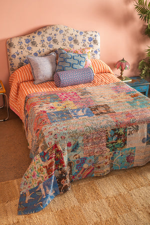 A bed with a floral headboard, orange striped sheets, and the Lola Patchwork Kantha Bed Throw by Ian Snow Ltd. Four patterned pillows are at the head, with a vintage pink lamp and leafy plant on a blue nightstand nearby.