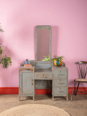The Grey Vintage Dresser Unit (W:105cm D:49cm H:166cm) by Ian Snow Ltd stands against a pink wall, featuring a tall mirror, faded blue paint, and multiple drawers. A plant in a colorful pot rests on top, with a chair and round rug nearby.