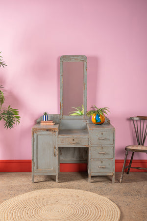 The Grey Vintage Dresser Unit (W:105cm D:49cm H:166cm) by Ian Snow Ltd stands against a pink wall, featuring a tall mirror, faded blue paint, and multiple drawers. A plant in a colorful pot rests on top, with a chair and round rug nearby.