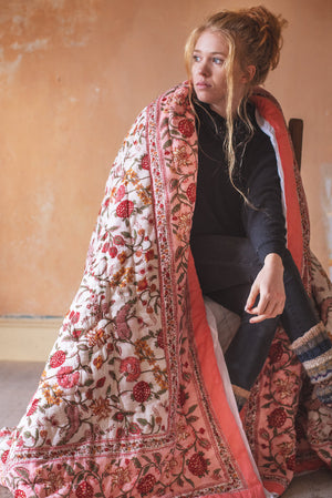 A woman with long, light brown hair sits on a chair, wrapped in the Coral Haze Reversible Floral Quilt by Ian Snow Ltd—its red, pink, and white floral patterns stand out in a softly lit rustic room with an orange wall.