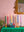 A row of colourful candles in Ian Snow Ltd’s Moulded Metal Ceramic Candle Holders sits on a green tablecloth, with old books and a gold ornate mirror against a pink wall. Touches of tableware and a plant leaf appear at the left.