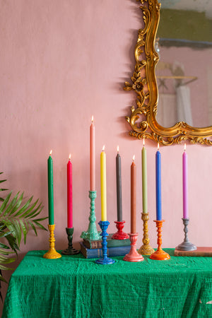 A row of colourful candles in Ian Snow Ltd’s Moulded Metal Ceramic Candle Holders sits on a green tablecloth, with old books and a gold ornate mirror against a pink wall. Touches of tableware and a plant leaf appear at the left.