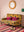 The Ian Snow Ltd Mustard Tree of Life Velvet Two Seater Sofa with two magenta cushions sits on a colorful striped rug. A round mirror hangs on the pale pink wall above, and a small side table with a green lamp completes the look.