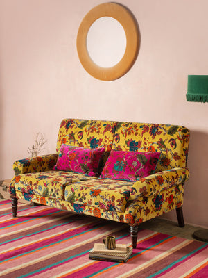 The Ian Snow Ltd Mustard Tree of Life Velvet Two Seater Sofa with two magenta cushions sits on a colorful striped rug. A round mirror hangs on the pale pink wall above, and a small side table with a green lamp completes the look.