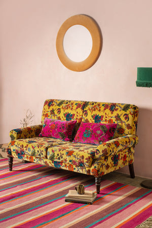 The Ian Snow Ltd Mustard Tree of Life Velvet Two Seater Sofa with two magenta cushions sits on a colorful striped rug. A round mirror hangs on the pale pink wall above, and a small side table with a green lamp completes the look.