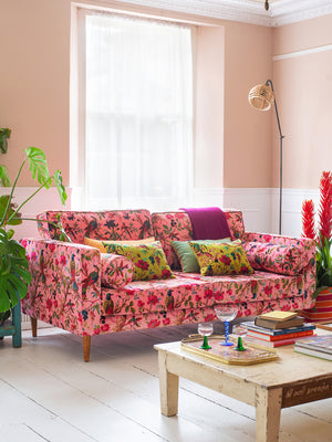 A bright living room with an Ian Snow Ltd Milla Pink Bird of Paradise Velvet Sofa, colorful cushions, a low wooden coffee table with books and glasses, light pink walls, a large window, and green and red potted plants.