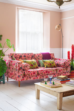 A bright living room with an Ian Snow Ltd Milla Pink Bird of Paradise Velvet Sofa, colorful cushions, a low wooden coffee table with books and glasses, light pink walls, a large window, and green and red potted plants.