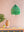 A Neon Green Paper Lampshade by Ian Snow Ltd hangs above a vibrant patterned table with a tan vase, small succulent, decorative box, and monstera leaf, all set against a pale pink wall.