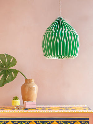 A Neon Green Paper Lampshade by Ian Snow Ltd hangs above a vibrant patterned table with a tan vase, small succulent, decorative box, and monstera leaf, all set against a pale pink wall.