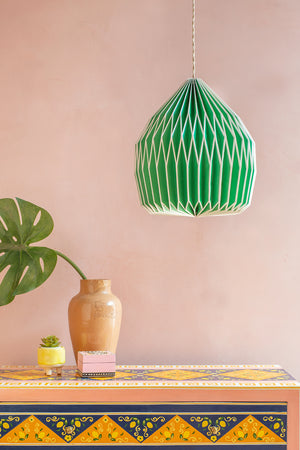 A Neon Green Paper Lampshade by Ian Snow Ltd hangs above a vibrant patterned table with a tan vase, small succulent, decorative box, and monstera leaf, all set against a pale pink wall.