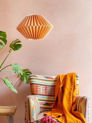 An Orange Paper Lampshade by Ian Snow Ltd with a geometric pattern hangs over an armchair with an orange blanket, while a monstera plant and wooden table sit nearby against a pale pink wall.