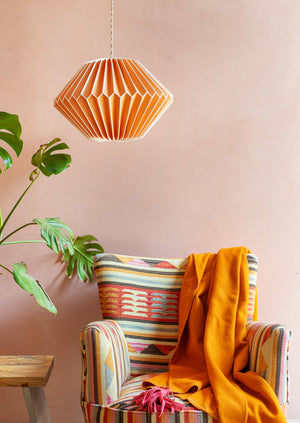 An Orange Paper Lampshade by Ian Snow Ltd with a geometric pattern hangs over an armchair with an orange blanket, while a monstera plant and wooden table sit nearby against a pale pink wall.