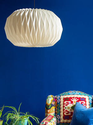 The Sphere Natural Pleated Paper Lampshade by Ian Snow Ltd hangs from the ceiling against a vibrant blue wall; below, a colorful patterned armchair and green potted plant complete the cozy room.