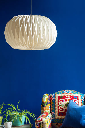 The Sphere Natural Pleated Paper Lampshade by Ian Snow Ltd hangs from the ceiling against a vibrant blue wall; below, a colorful patterned armchair and green potted plant complete the cozy room.