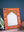 The Orange Hand Painted Arched Wooden Mirror by Ian Snow Ltd features an ornate orange and red floral frame, resting against a dark blue wall on a light wood surface, with green plant leaves and décor nearby.