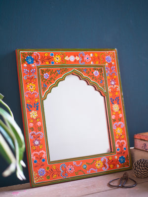 The Orange Hand Painted Arched Wooden Mirror by Ian Snow Ltd features an ornate orange and red floral frame, resting against a dark blue wall on a light wood surface, with green plant leaves and décor nearby.