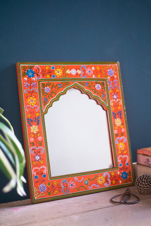The Orange Hand Painted Arched Wooden Mirror by Ian Snow Ltd features an ornate orange and red floral frame, resting against a dark blue wall on a light wood surface, with green plant leaves and décor nearby.