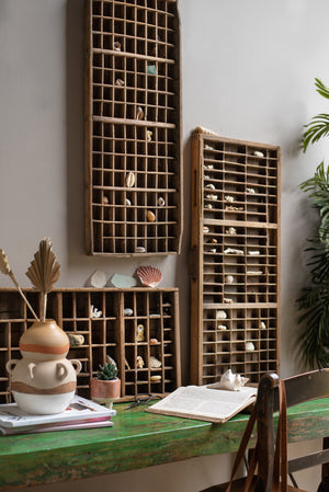An eclectic workspace featuring the Vintage Wooden Printing Press Tray by Ian Snow Ltd filled with shells and trinkets, a green rustic desk, decorative ceramics, an open book, and a potted plant in the background.